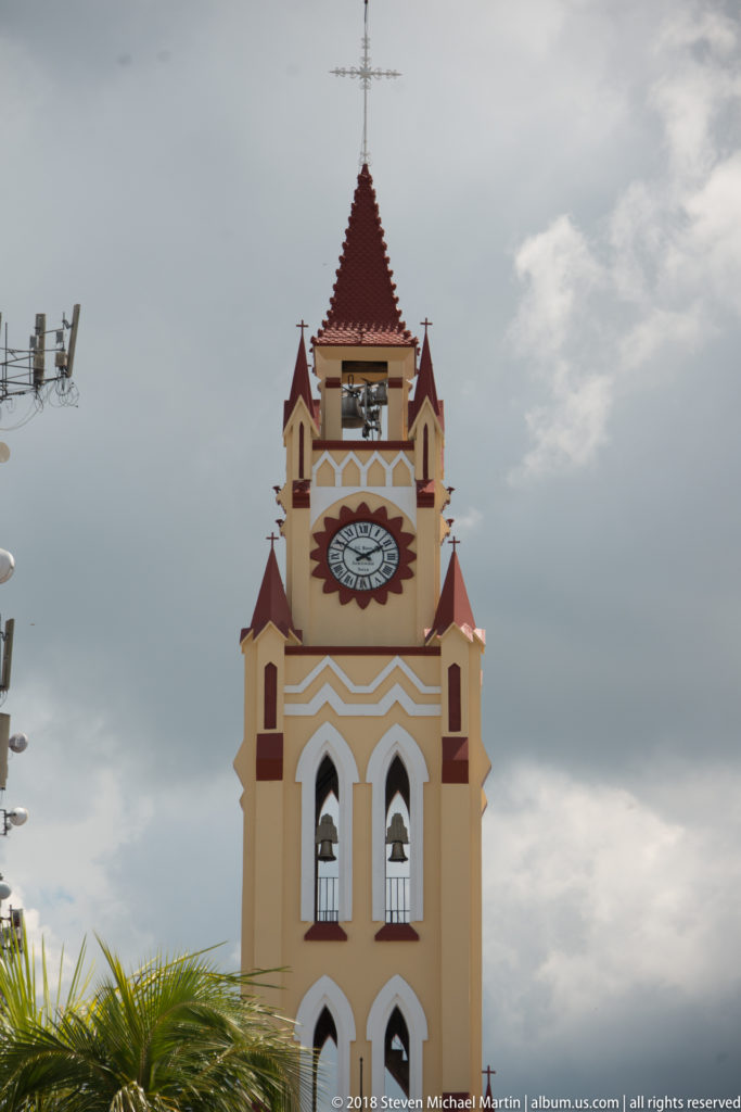 Upper clock tower of Iquitos Cathedral with red trim, arched openings, steep roof, and cross.