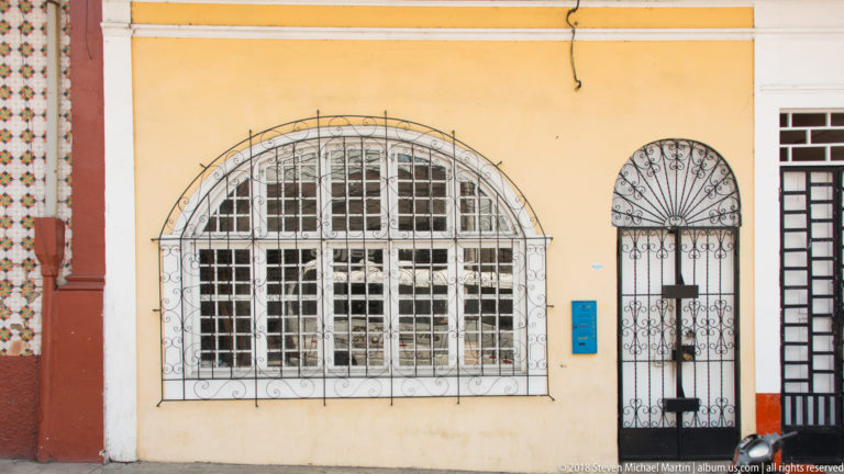Yellow façade with arched window and narrow doorway covered by decorative white and black iron grilles.