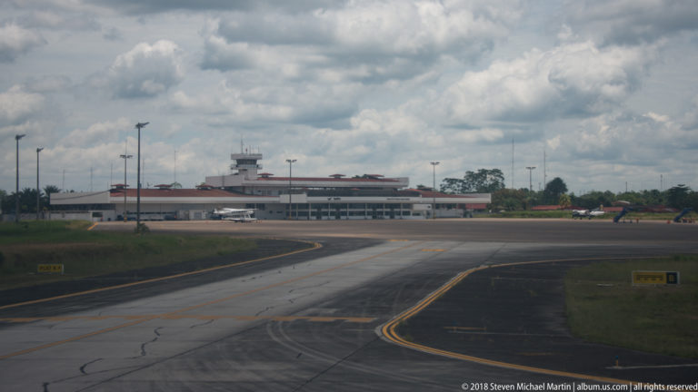 Airport terminal buildings and tower-like structure beyond taxiways under a cloudy sky.