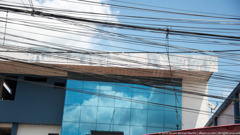 Blue-glass modern building crossed by dense black overhead utility wires against a bright Iquitos sky.