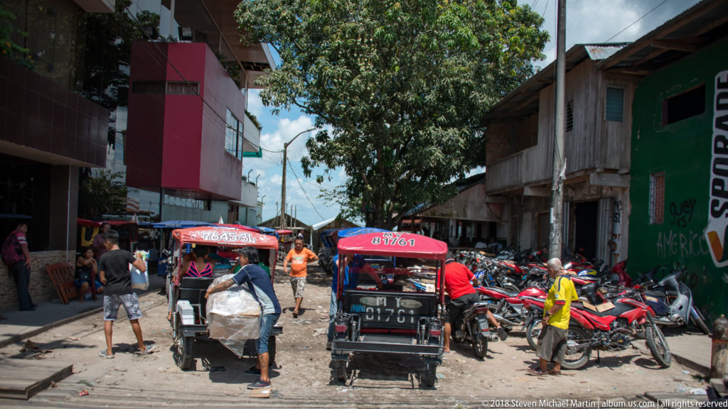 Mototaxis, motorcycles, and pedestrians crowd a narrow sunlit street lined with weathered Iquitos buildings.