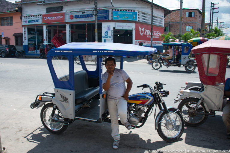 Man standing beside a blue-canopied Honda mototaxi on a bright Iquitos street.