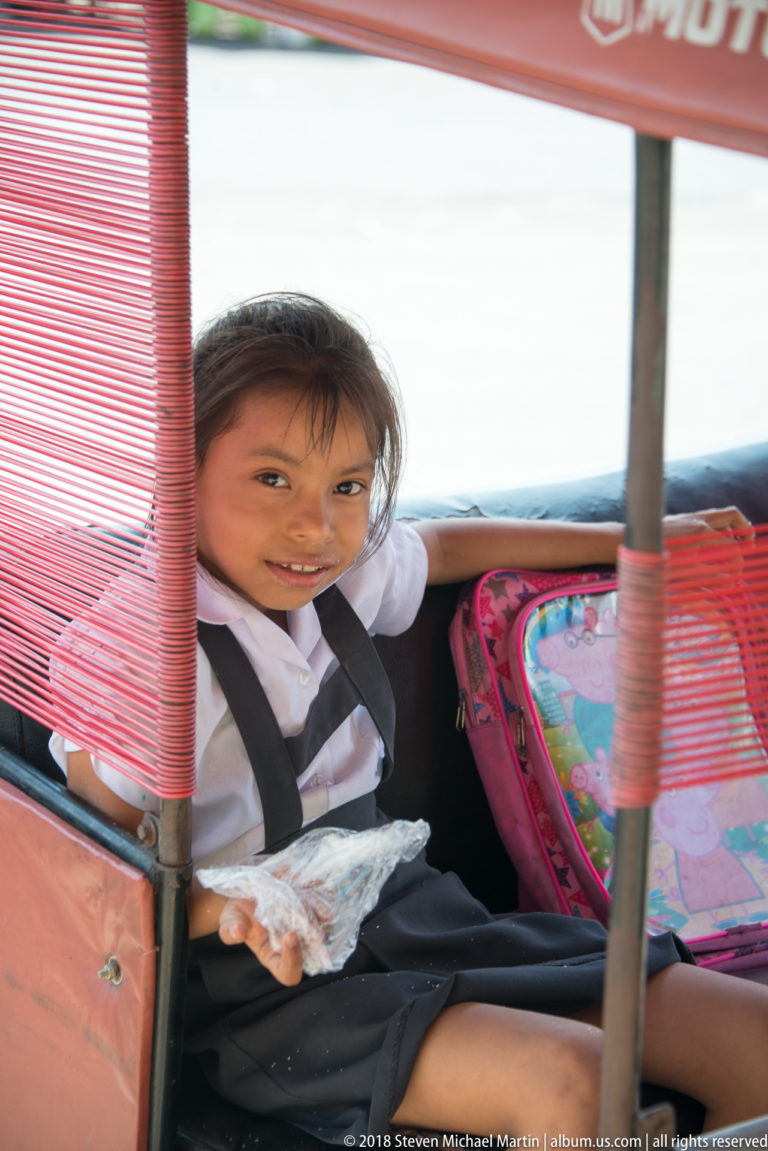 Smiling schoolgirl in a mototaxi passenger seat with a colorful backpack beside her.