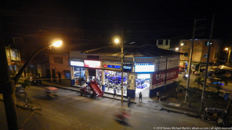 Nighttime Iquitos intersection with illuminated storefronts, streetlights, pedestrians, and blurred traffic.