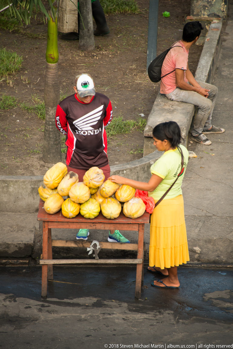 Vendor behind a wooden table piled with large yellow produce as a woman stands beside the stall.