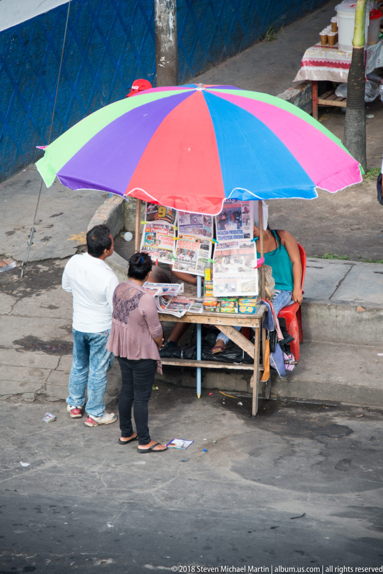 Sidewalk newsstand covered by a multicolored umbrella, with printed publications displayed around the kiosk.