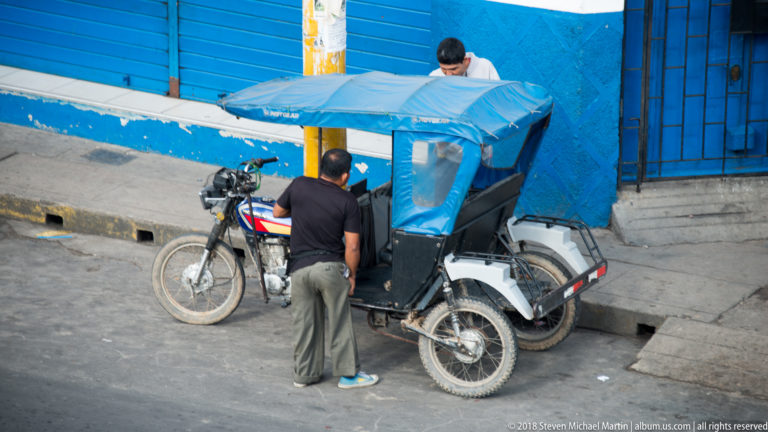 Man beside a blue-canopied three-wheeled mototaxi parked against a bright blue wall in Iquitos.