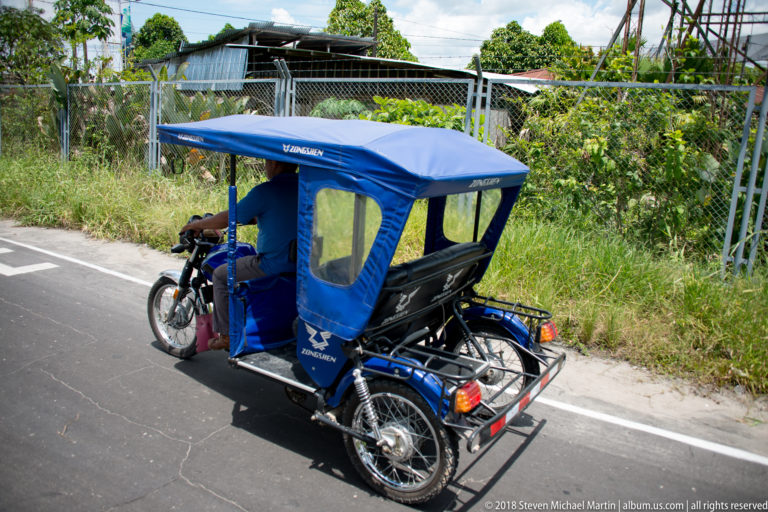 Rear view of a blue mototaxi traveling beside grass and chain-link fencing.