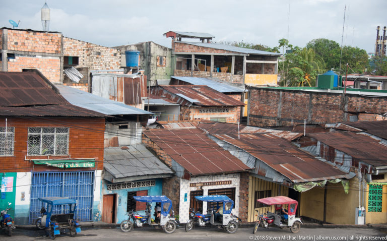 Elevated Iquitos block with rusted corrugated roofs, unfinished brick buildings, storefronts, and parked mototaxis.