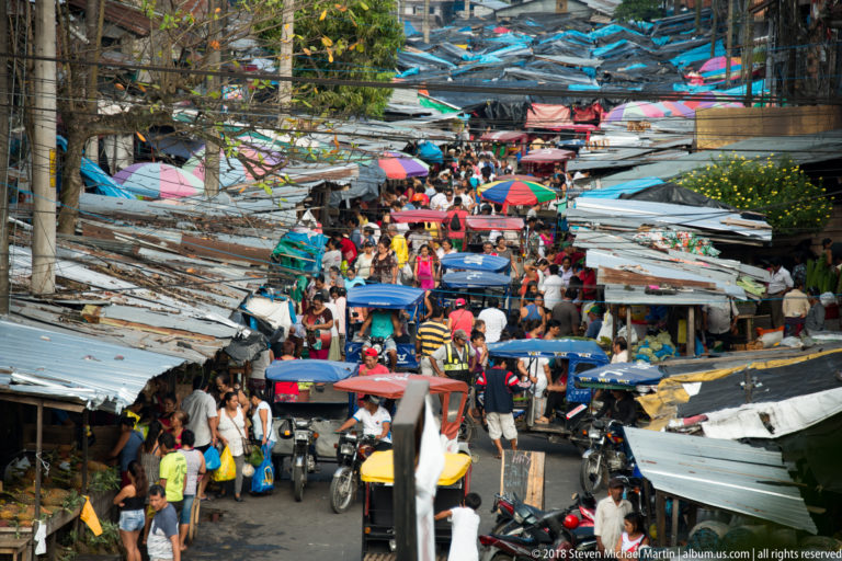 Crowded Iquitos market street filled with pedestrians, mototaxis, motorcycles, stalls, and colorful umbrellas.