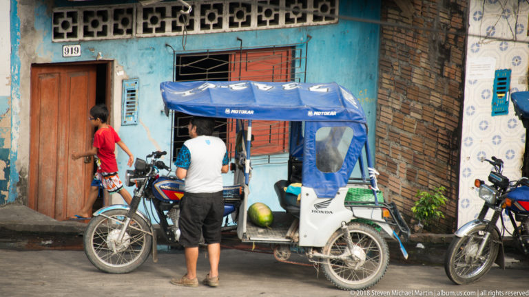 Man standing beside a blue-canopied mototaxi near a doorway and another motorcycle in Iquitos.
