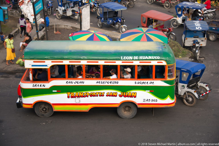 Green Transportes San Juan bus marked with Iquitos route names amid mototaxis and market umbrellas.