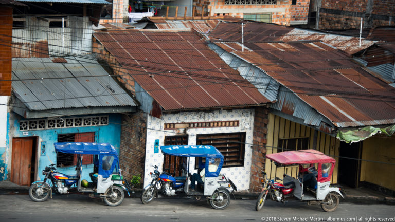 Three covered mototaxis parked before narrow storefronts with rusted corrugated roofs in Iquitos.
