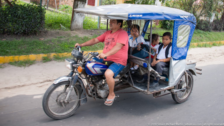 Blue-and-white mototaxi carrying several school-age passengers along an Iquitos street.
