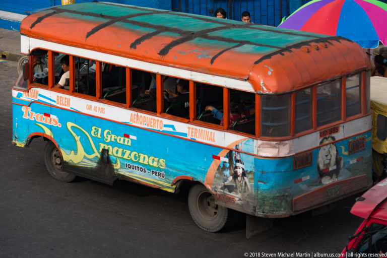 Weathered turquoise-and-orange El Gran Amazonas bus carrying passengers on an Iquitos airport route.