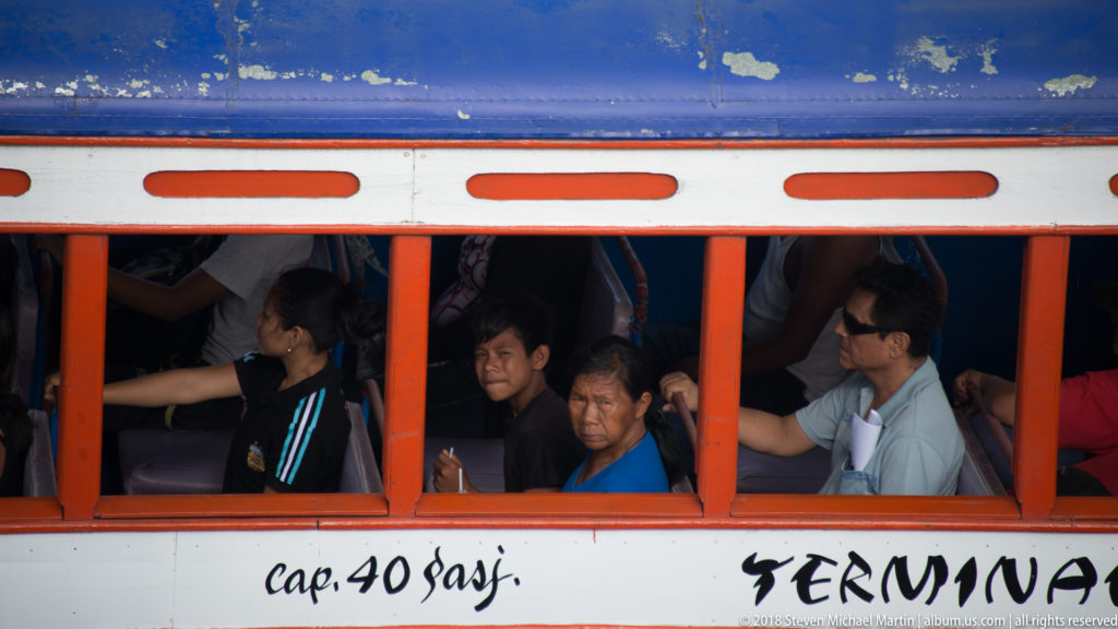 Passengers seated behind orange-framed open windows of a crowded local bus in Iquitos.
