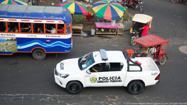 White Peruvian National Police pickup among a bus, mototaxis, and colorful market umbrellas in Iquitos.