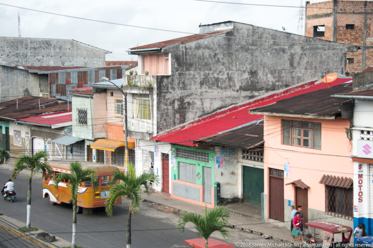 Elevated view of low-rise Iquitos buildings with corrugated roofs, palms, overhead wires, and a bus on the street.