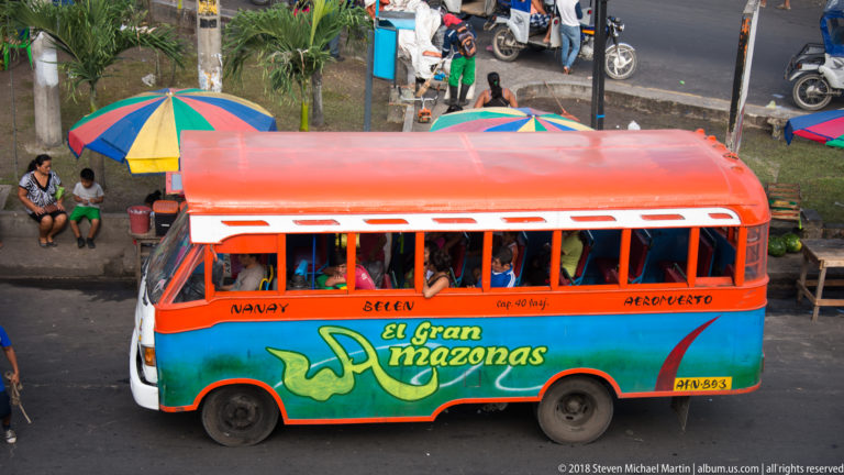 Colorful El Gran Amazonas bus with passengers passing curbside vendors and umbrellas on an Iquitos street.