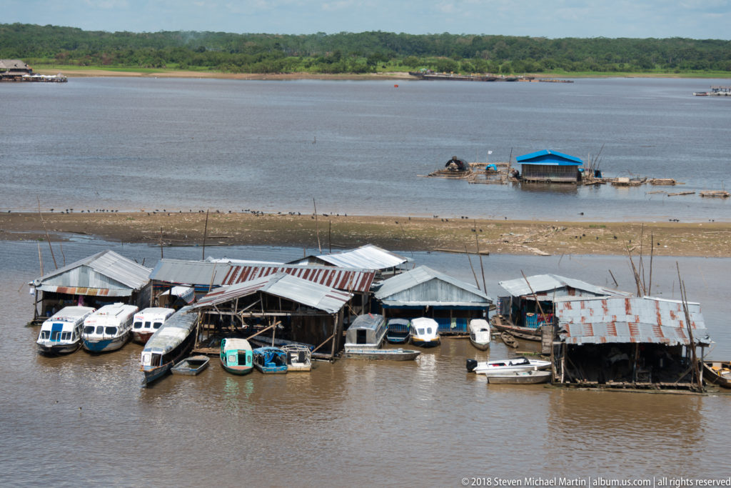 Elevated view of corrugated-roof dock structures, several enclosed passenger launches, mudflats, and a blue-roofed floating building.