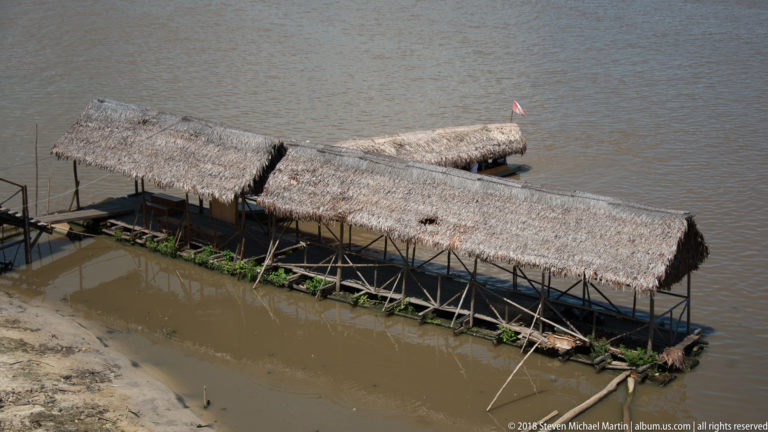 Long floating platform with several palm-thatch roofs and open timber framing beside a muddy riverbank.