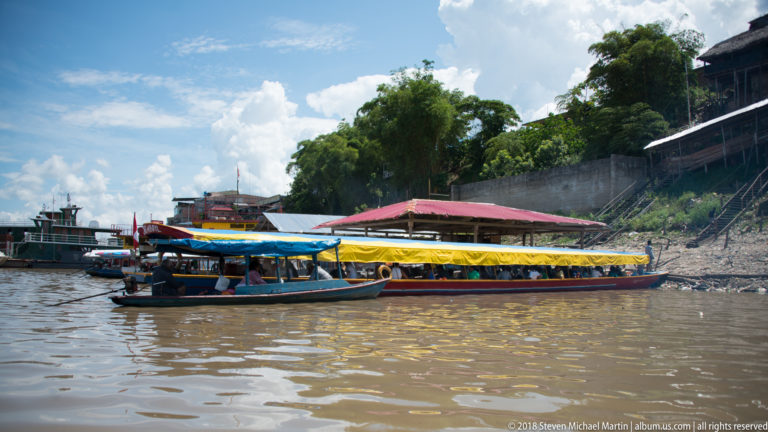 Two long passenger motorboats with blue and yellow canopies moored beside a red-roofed riverside landing.