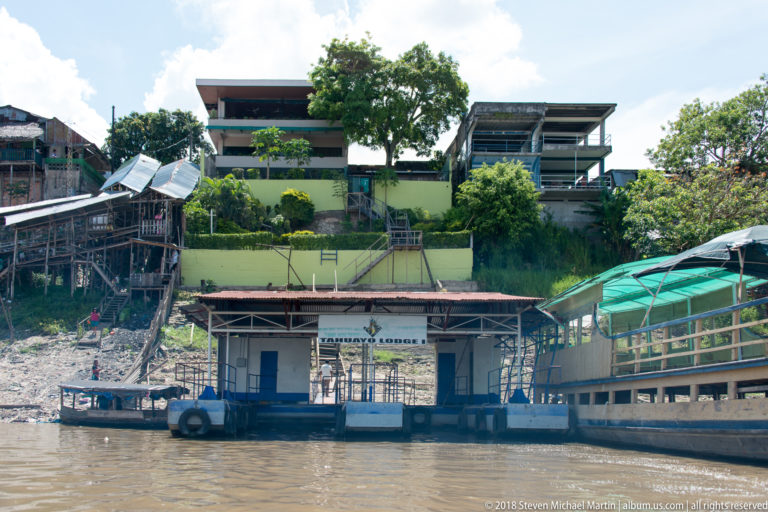 Small floating dock building with painted signage and tire fenders below a steep developed riverbank.