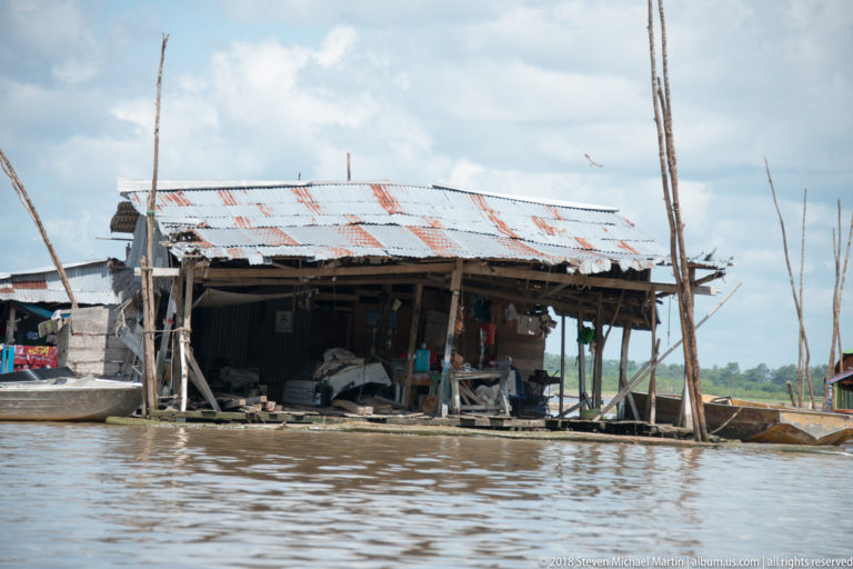 Open timber shed with rusted corrugated roof at the river edge, with small boats and stored materials around it.