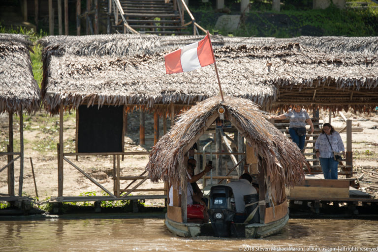 Small motorboat beneath a palm-thatch canopy at a timber landing with a Peruvian flag overhead.