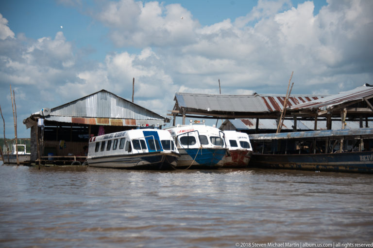 Three enclosed white and blue passenger launches moored beside a corrugated-roof shed on the Iquitos waterfront.