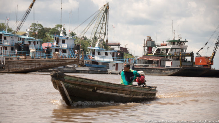 Small wooden motor canoe carrying two people and cargo across brown water in front of larger docked vessels.