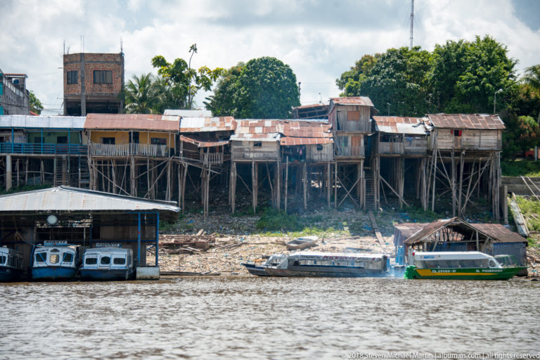 Weathered wooden houses on tall stilts above a muddy riverbank with small motorboats moored below.