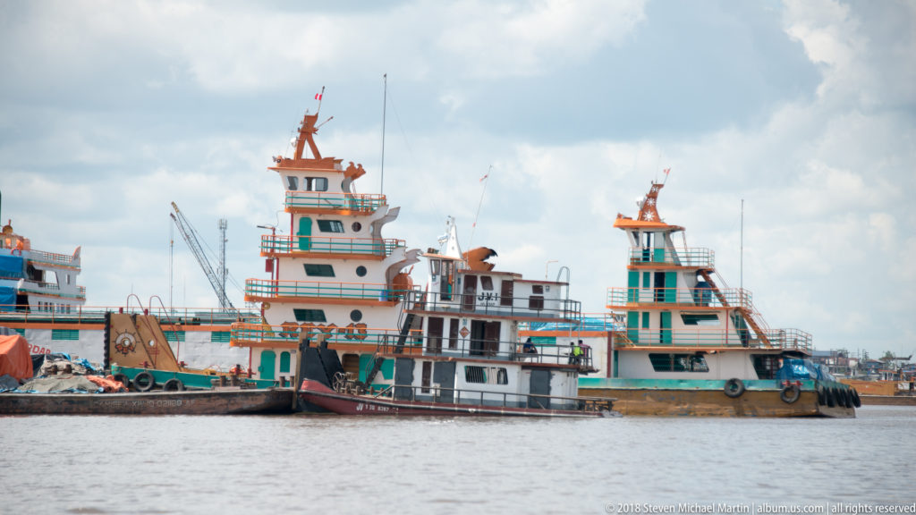 Several decorated workboats and smaller craft moored along the Iquitos waterfront beneath a cloudy tropical sky.