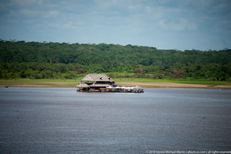 Isolated floating structure with broad palm-thatch roofs on the river before a densely forested green bank.