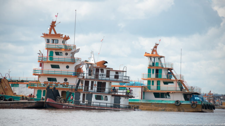 Three colorful workboats with tall wheelhouses, turquoise railings, and tire fenders moored together on brown river water.