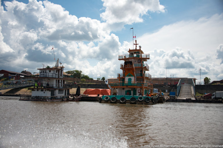 Orange river workboat with tall wheelhouse and tire fenders moored beside a sloping landing under dramatic clouds.