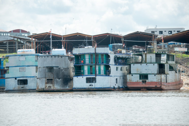 Weathered multi-deck riverboats moored closely beside large open-sided sheds on the Iquitos waterfront.