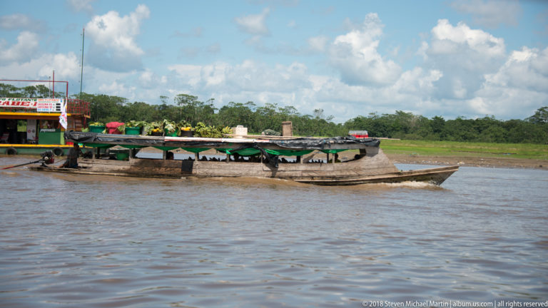 Low elongated wooden riverboat with dark canopy traveling on brown water near a green riverbank.