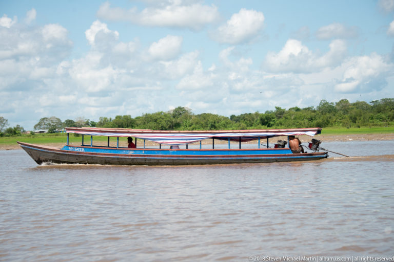 Long brightly painted covered canoe crossing brown river water with a driver seated at the stern.