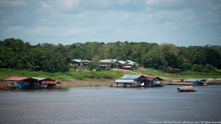 Distant riverside houses, floating buildings, and small boats along a forested Amazon bank across broad water.
