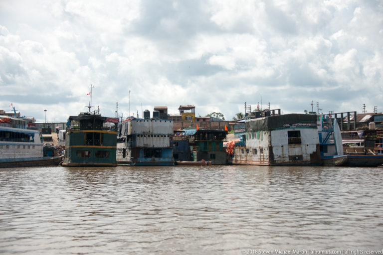 Several large, weathered riverboats moored side by side along the Iquitos waterfront under a cloudy sky.