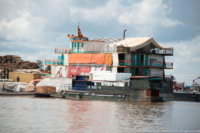 Large multi-deck riverboat with turquoise railings, tarps, and advertising banners moored along the Iquitos waterfront.