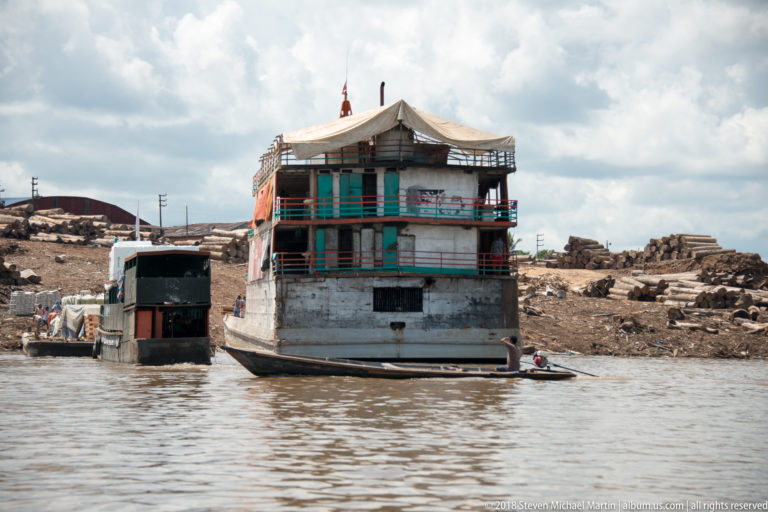 Large weathered multi-deck riverboat moored beside a rough shoreline with stacked material and small craft nearby.