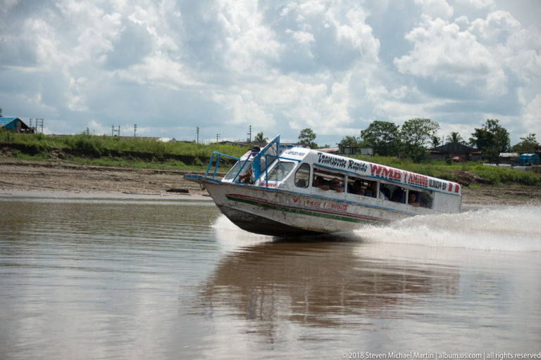 Covered passenger speedboat skimming across brown river water with bow raised and white spray trailing behind.