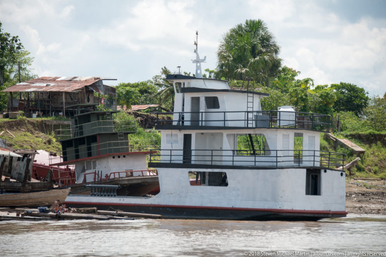 White multi-level river vessel beside other boats at a muddy riverside repair or construction area.