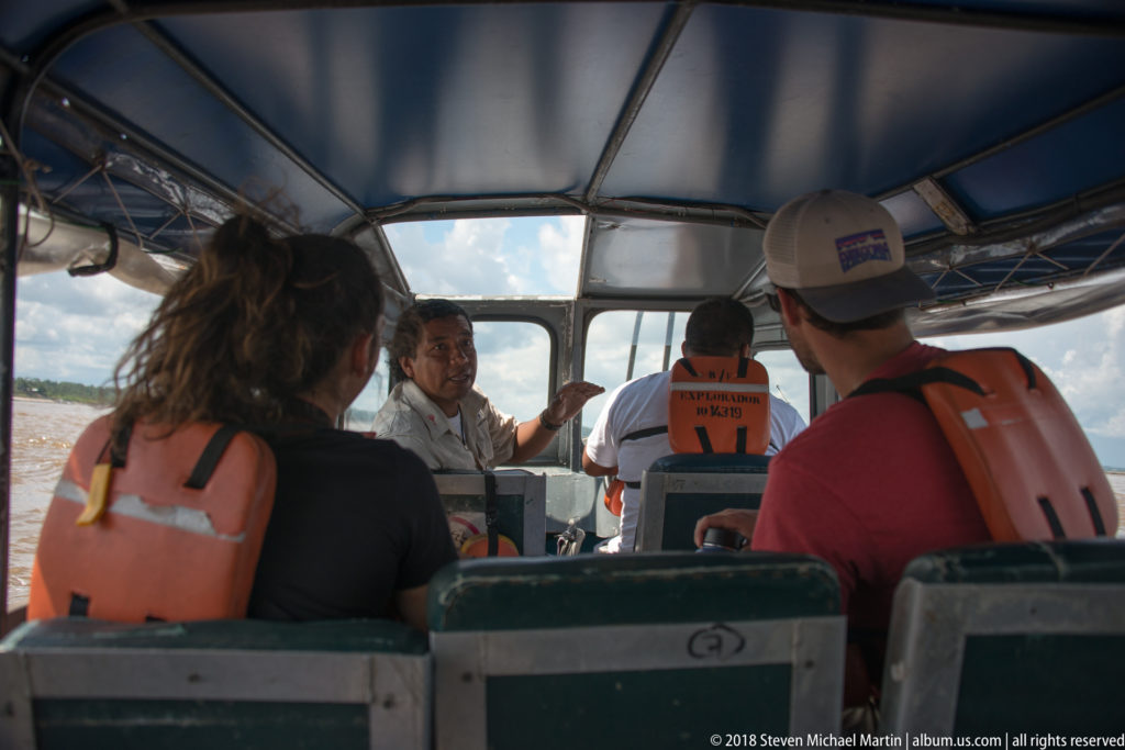 Rear view inside a covered river launch with passengers in life jackets and a crew member gesturing near the helm.