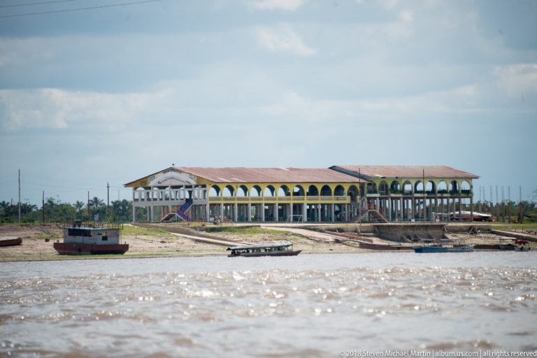Large arcaded open-sided building above a muddy river landing with several small boats along the shore.