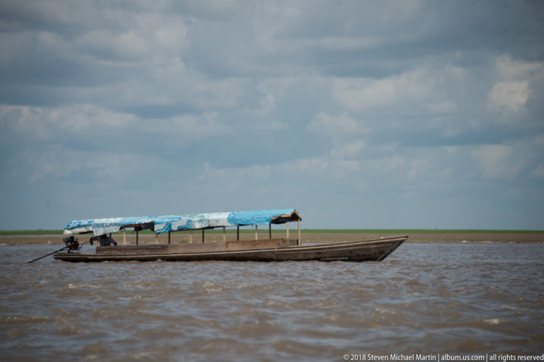 Long narrow motor canoe with blue canopy traveling across choppy brown river water under gray clouds.