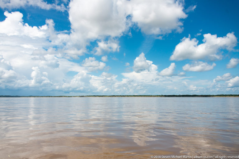 Wide brown river under towering white clouds with a low, distant green shoreline near Iquitos.