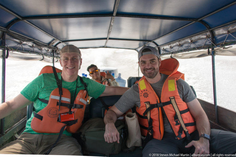 Two male passengers in orange life jackets seated beneath a blue canopy inside an open-sided river launch.